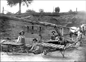 Figure 2. Close behind the pug mill is the excavated face of a clay bank. Tempered clay was taken from the mill and put into a wood box, shown next to the kneeling man. From there it was put into molds, which can be seen on the barrows.