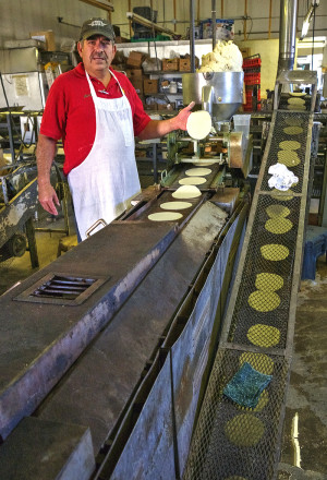 Martin Palma holds a perfectly formed raw tortilla that has just come out of the hopper. Tortillas are seen traveling along the conveyor belt system; in, through and out of the three level oven in the foreground. Photo by John McEvoy.